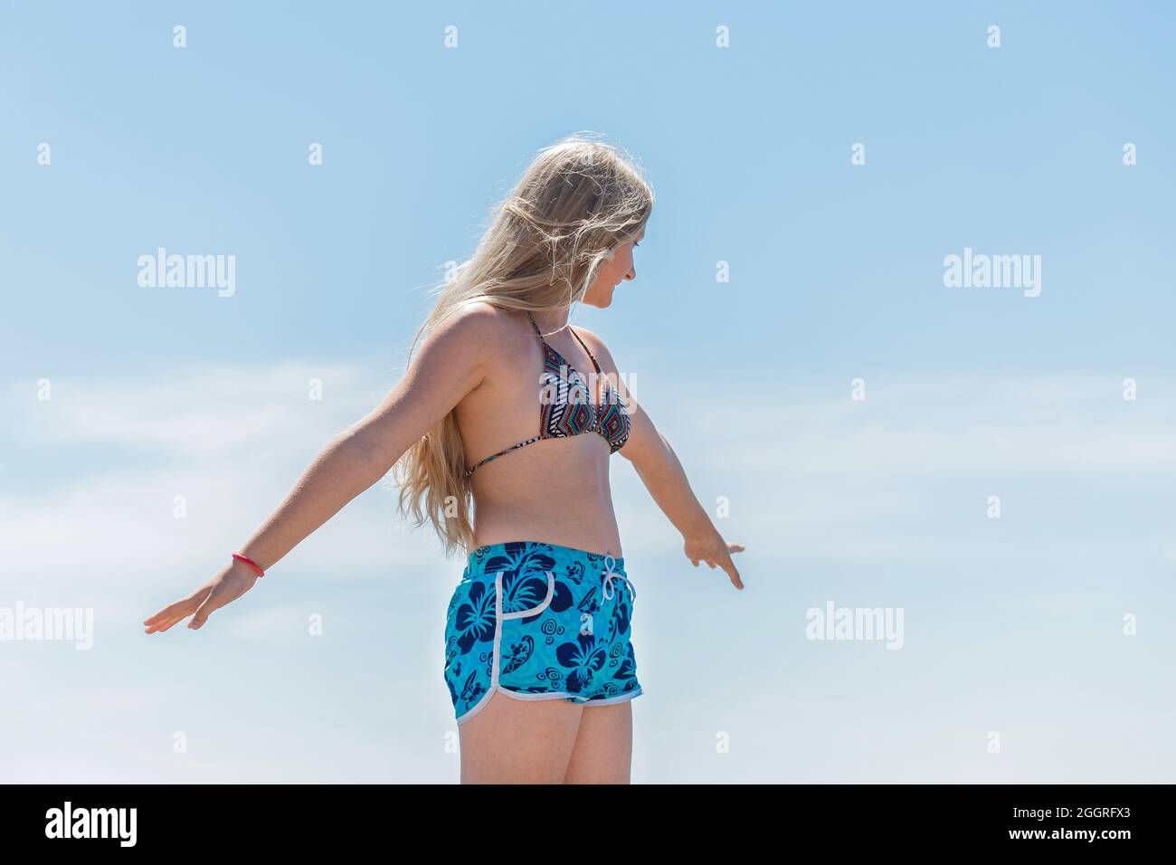Ein junges blondes Mädchen, ein Teenager mit europäischem Auftritt in einem Badeanzug und Shorts, posiert mit den Händen an den Seiten gegen den blauen Himmel. Stockfoto