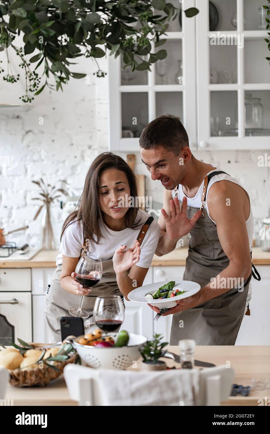 Glückliches Paar, das Videoanruf mit dem Smartphone macht, während es das Abendessen in der Küche zu Hause zubereitet fröhliche kaukasische Menschen winken ihre Hände und lächeln Stockfoto