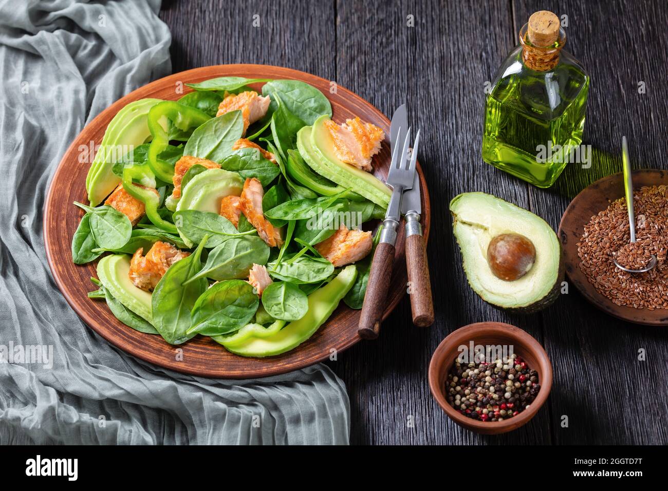 Lachs Spinat Avocado und grüner Pfeffer Salat auf einem braunen rustikalen Teller mit Gabel und Messer auf einem Mooreichentisch, mediterrane gesunde Ernährung Stockfoto