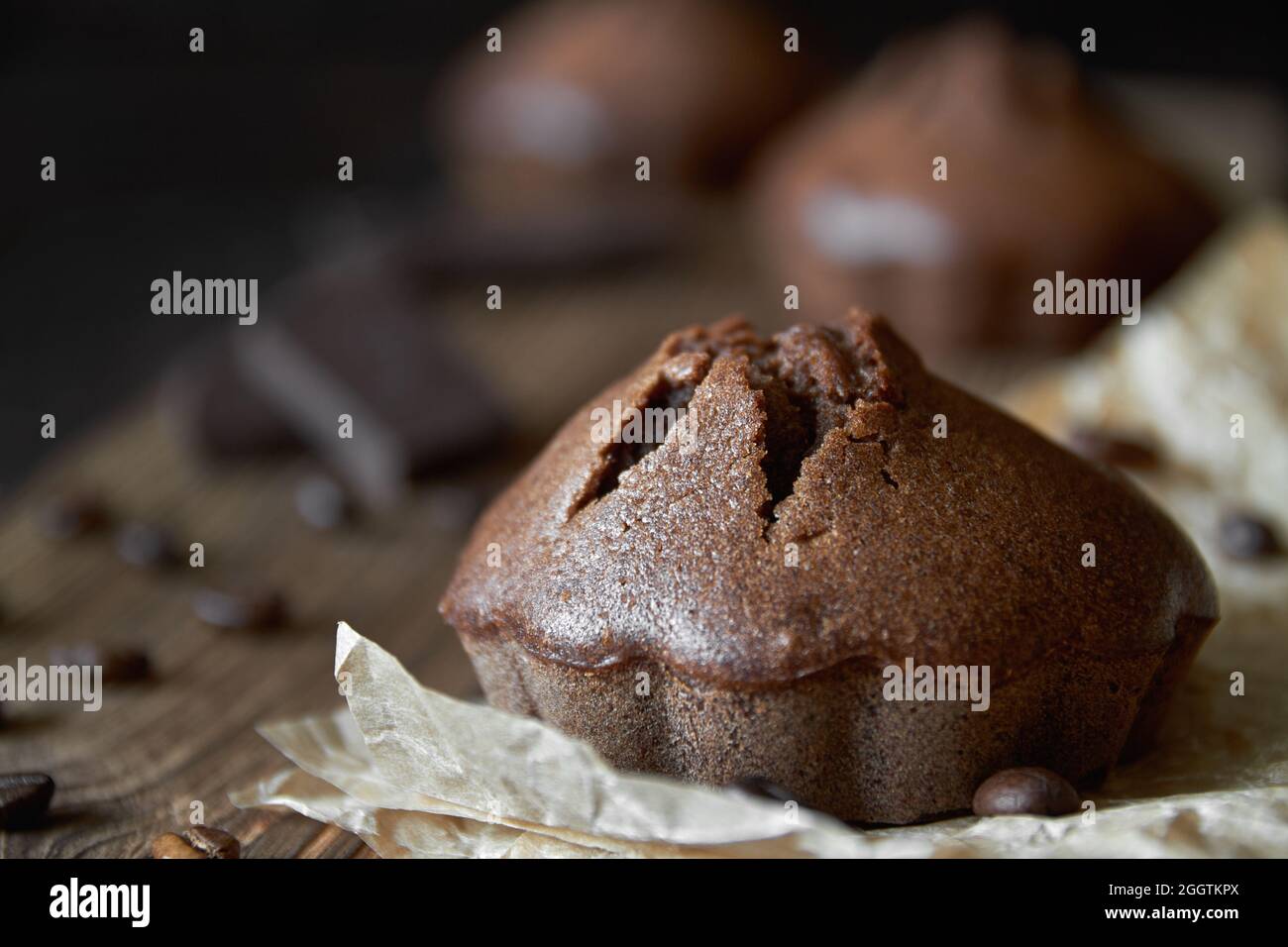 Hausgemachte Schokoladen-Muffins auf Pergament mit Kaffeebohnen und Schokoladenstücken. Dunkler Holzhintergrund, Nahaufnahme, selektiver Fokus Stockfoto