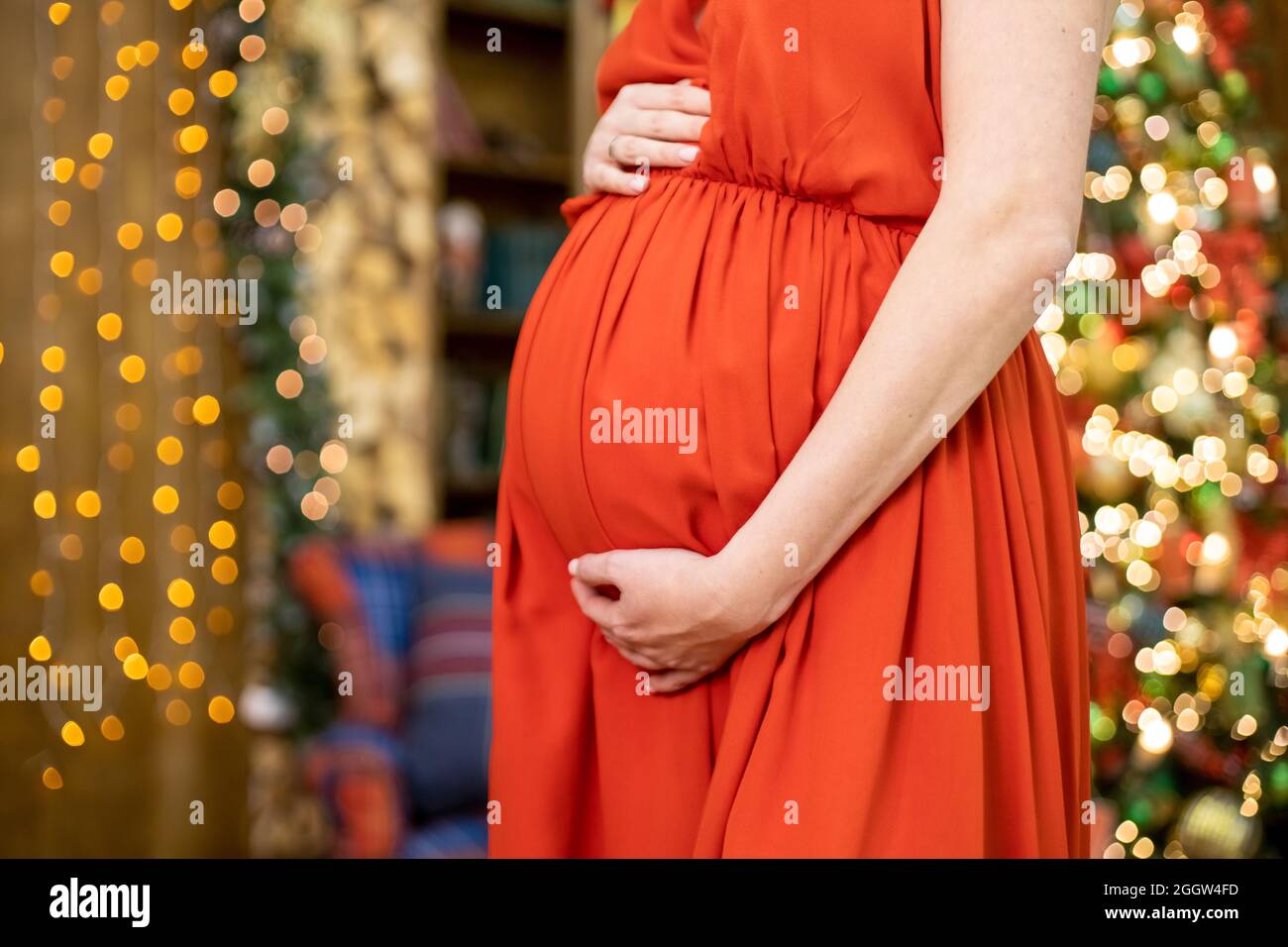 Bestes weihnachtsgeschenk. Gesichtslose Schwangere in einem roten Kleid umarmt ihren Bauch vor dem Hintergrund des Weihnachtswohnzimmers in Unschärfe. Nahaufnahme kein fa Stockfoto