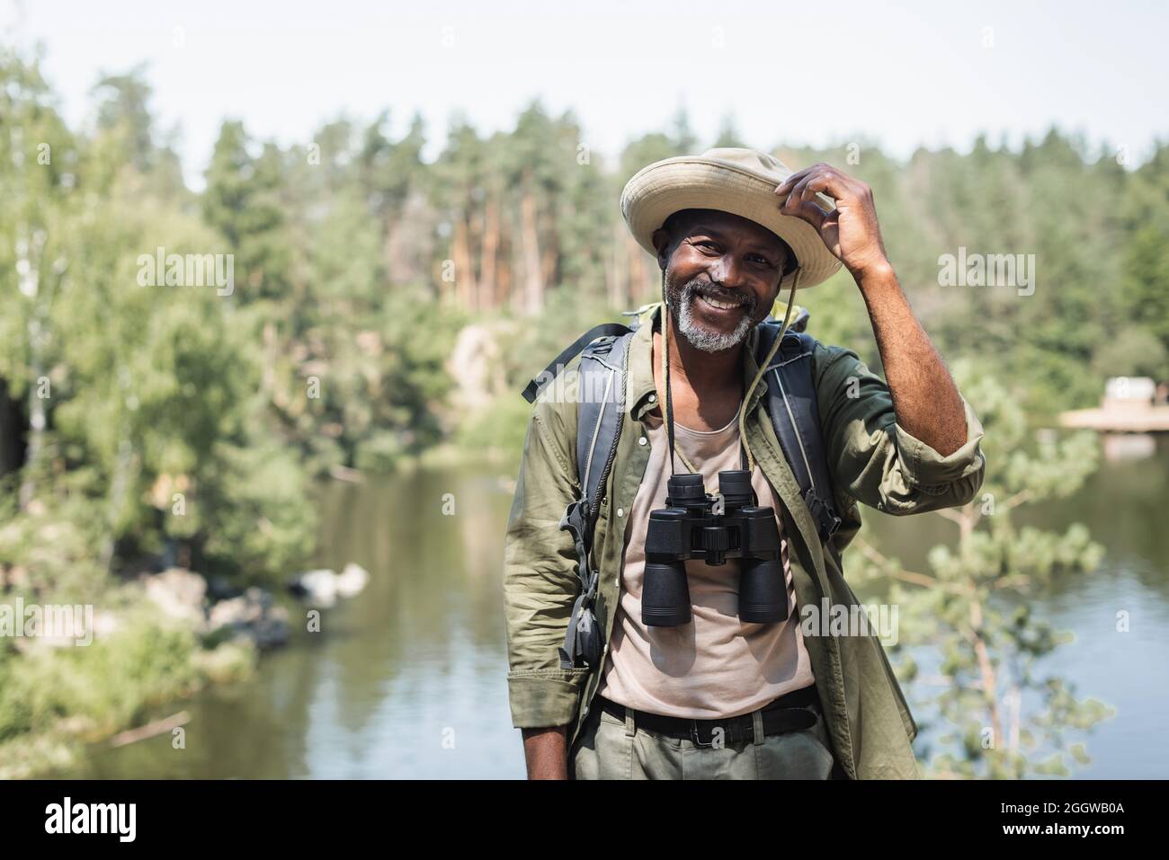 Positiver afroamerikanischer Wanderer mit Hut und Fernglas, der im Freien auf die Kamera schaut Stockfoto