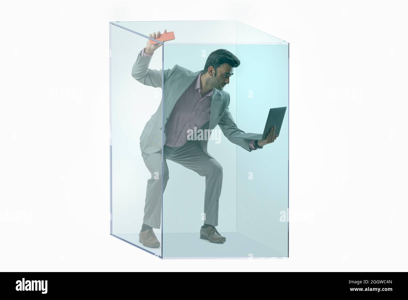 Porträt eines Mannes in formellem Business-Anzug mit Laptop und Handy in einem durchsichtigen Würfel. Stockfoto