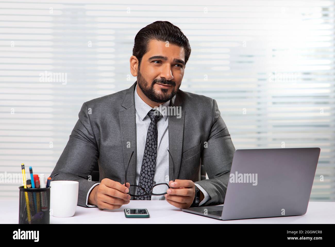 Ein Mitarbeiter, der im Büro sitzt und mit einem Laptop eine Brille hält, schaut sich woanders um. Stockfoto