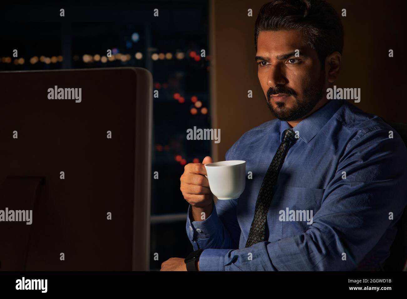 Ein Mitarbeiter hält einen Kaffeebecher, der nachts mit intensivem Blick im Büro sitzt. Stockfoto