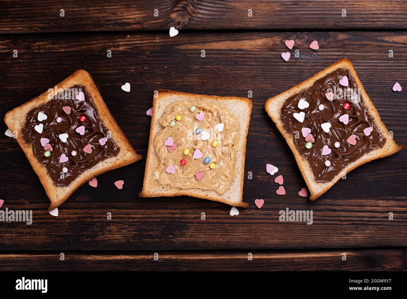 Drei Toast mit Schokoladenpaste und Erdnussbutter, mit bunten Herzen bestreut, auf einem dunklen Holzhintergrund, Draufsicht. Stockfoto