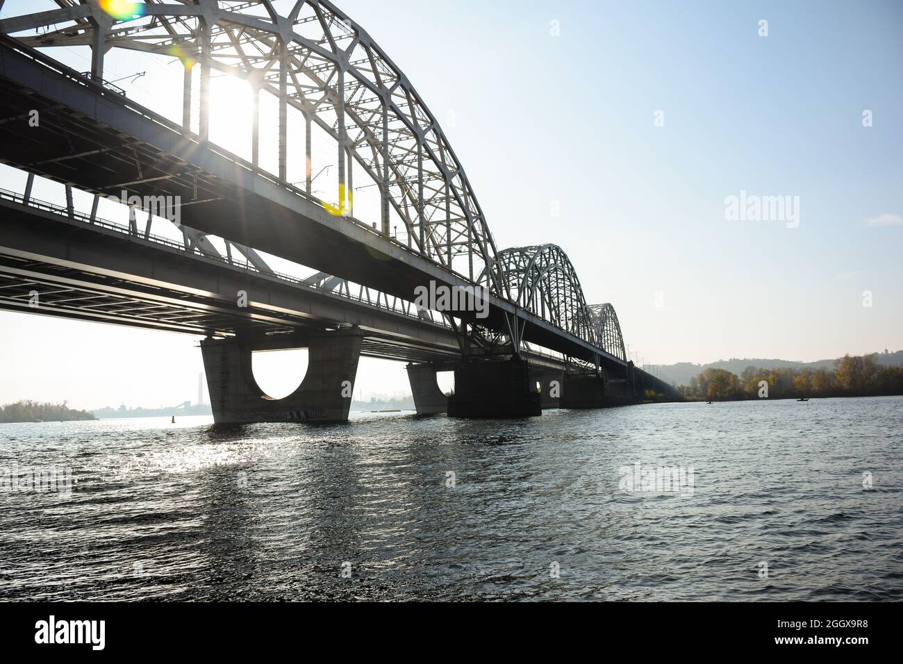Lange gewölbte Eisenbrücke über einen breiten Fluss. Sonnenstrahlen durchdringen sie. Vor dem Hintergrund des blauen Himmels. Stockfoto