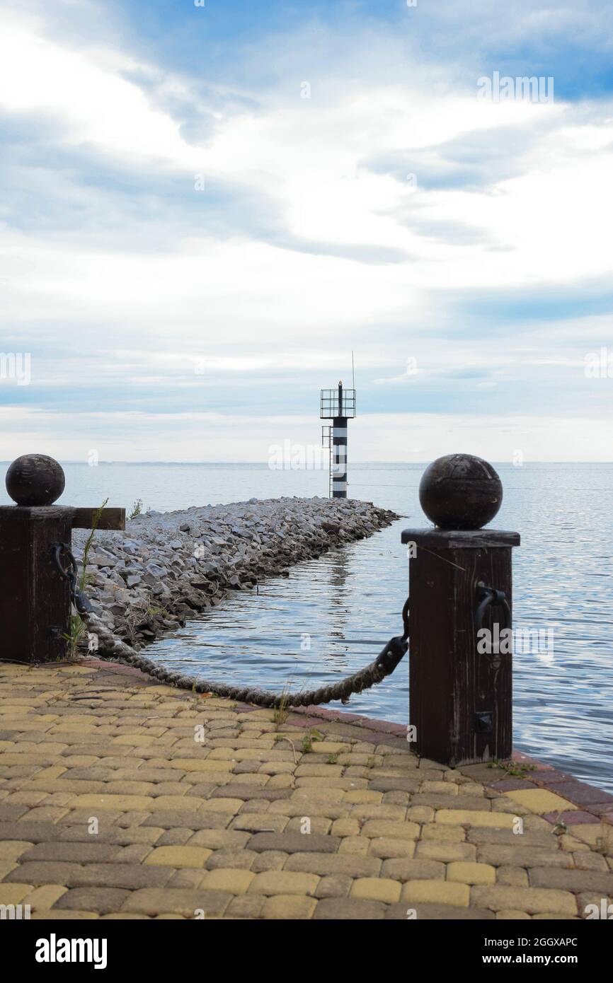 Alte felsige Pier mit Holzpfosten, ein Leuchtturm auf einem Steindamm im Hintergrund und ein heller Himmel Stockfoto