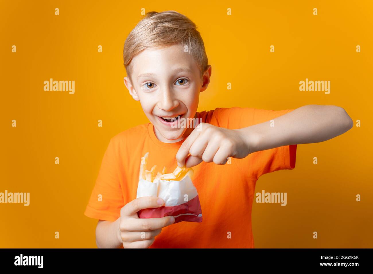 Ein Junge in einem orangefarbenen T-Shirt hält eine Tüte frittierter Kartoffeln vor sich Stockfoto