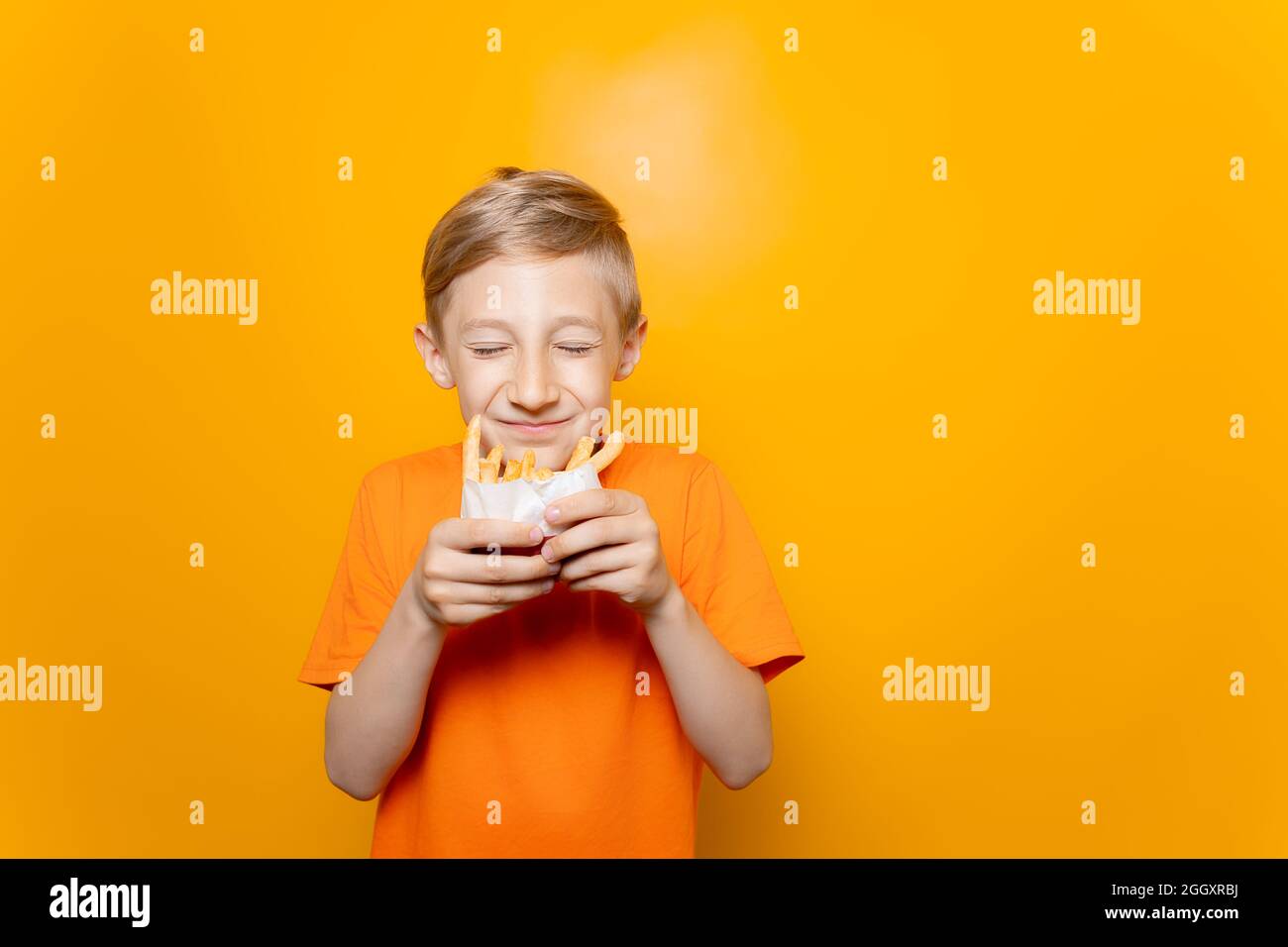 Ein Junge in einem orangefarbenen T-Shirt hält eine Tüte frittierter Kartoffeln vor sich und schnüffelt sie mit geschlossenen Augen Stockfoto