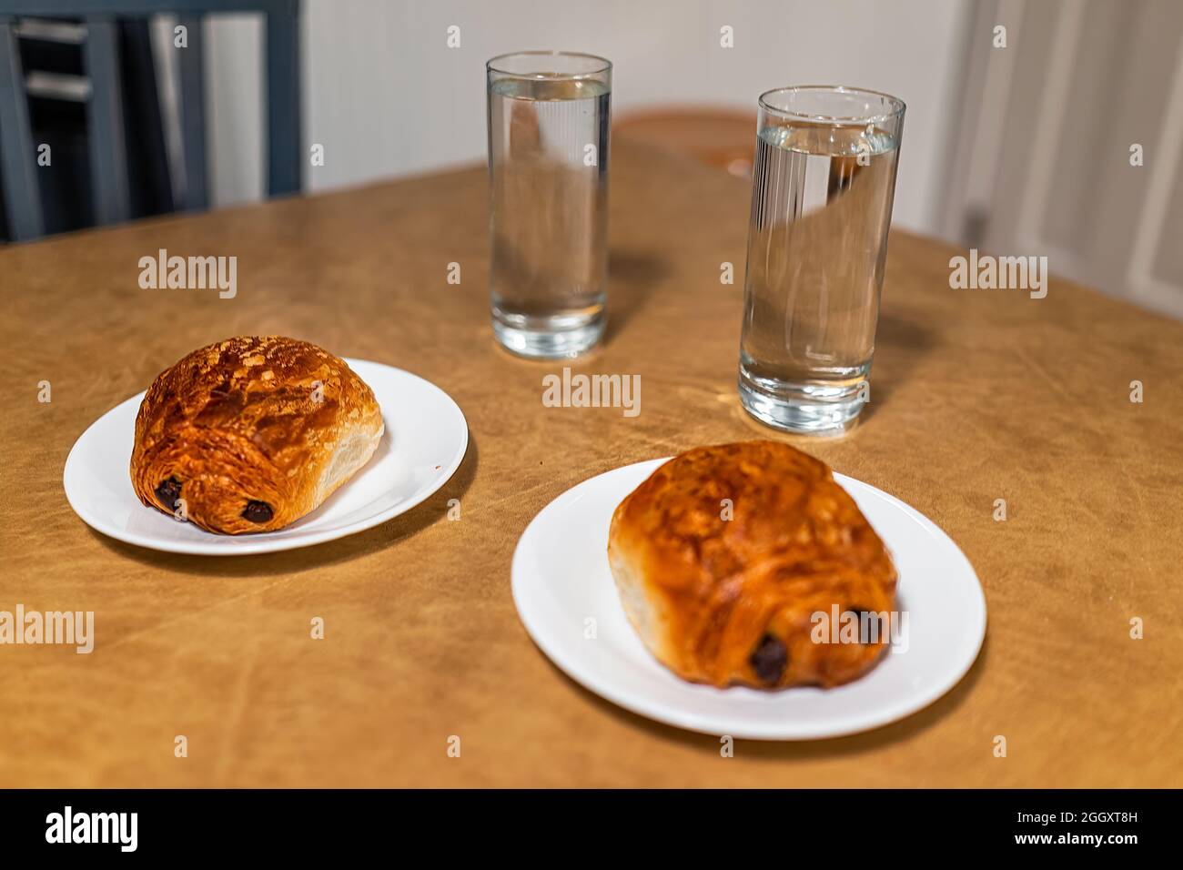 Zwei goldbraun gebackene Croissants auf Tellern mit schuppigen Krusten und Schokoladenfüllung mit Gläsern Wasser zum Frühstück Stockfoto