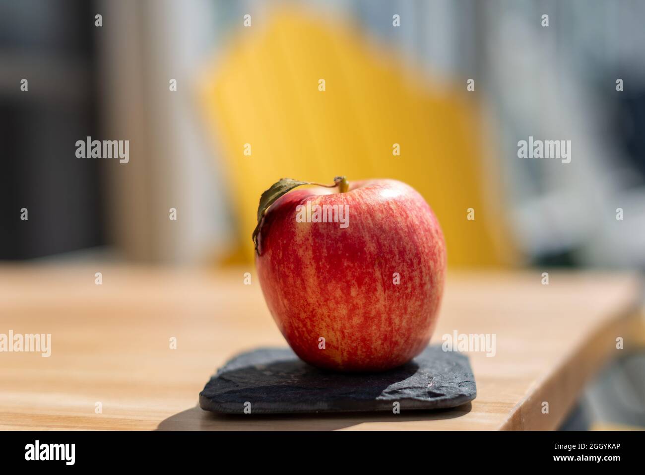 Ein roter, gala, Apfel sitzt auf einem kleinen quadratischen grauen Schiefer auf einem Holztisch. Die süße, knackige Frucht hat eine feste und glänzende Haut mit einer lebensmitteltauglichen Wachsbeschichtung. Stockfoto