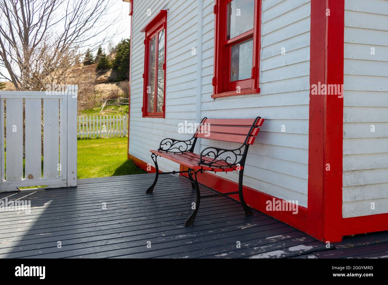 Die Außenwand eines weißen Hauses mit rot getrimmten Doppelfenstern, eine rote Holzbank mit schwarzen Eisenbeinen auf einer grauen Holzterrasse. Stockfoto