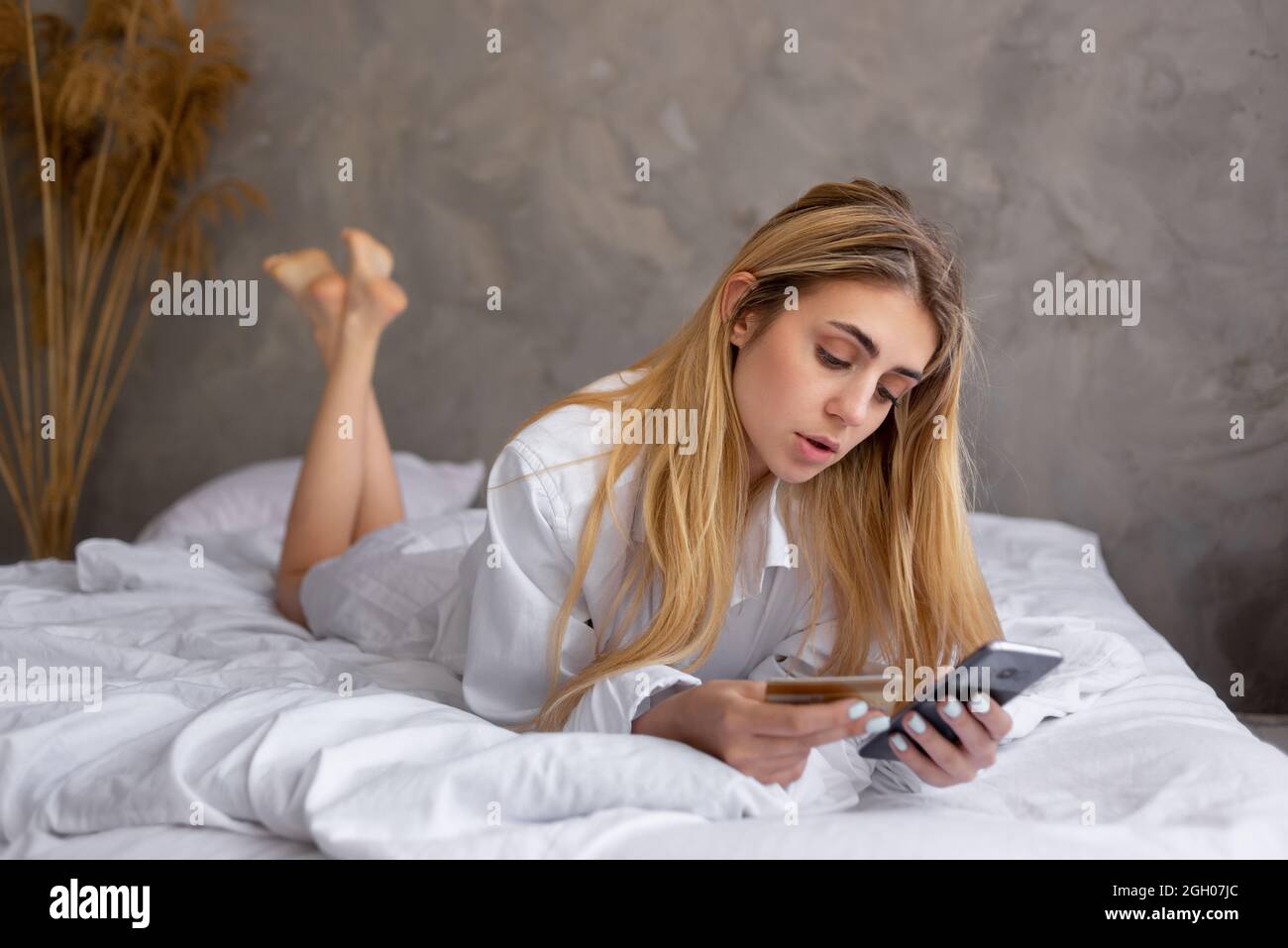 Porträt eines jungen schönen kaukasischen Mädchen auf dem Bett zu Hause liegen, Online-Shopping. Verwendet eine Kreditkarte und sein Smartphone. Stockfoto