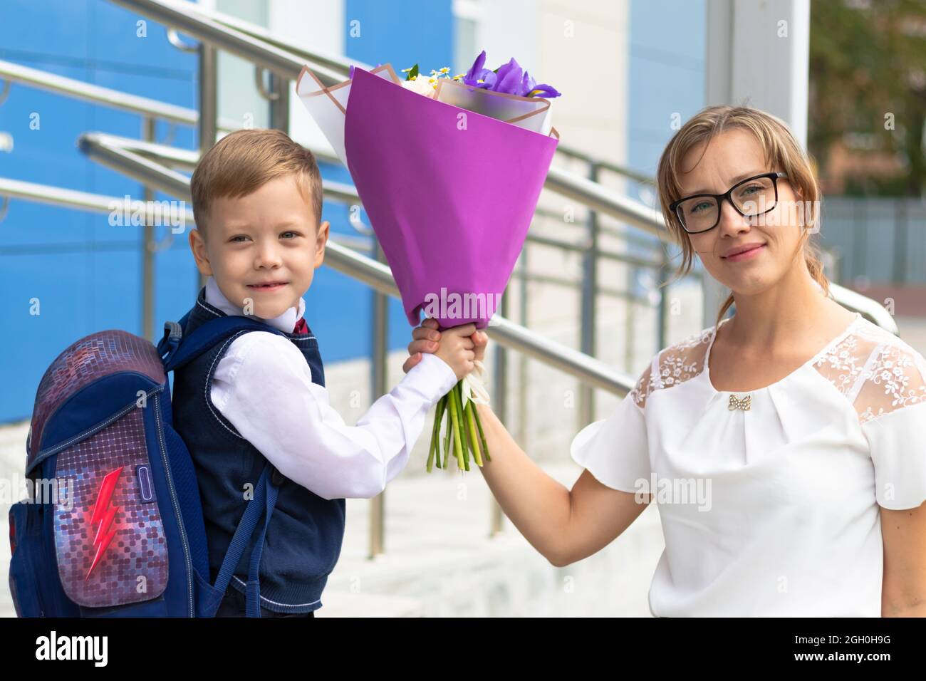 Ein süßer Erstklässler-Junge in Schuluniform mit seiner Mutter und einem schönen Blumenstrauß im Schulhof. Feier am 1. September Stockfoto