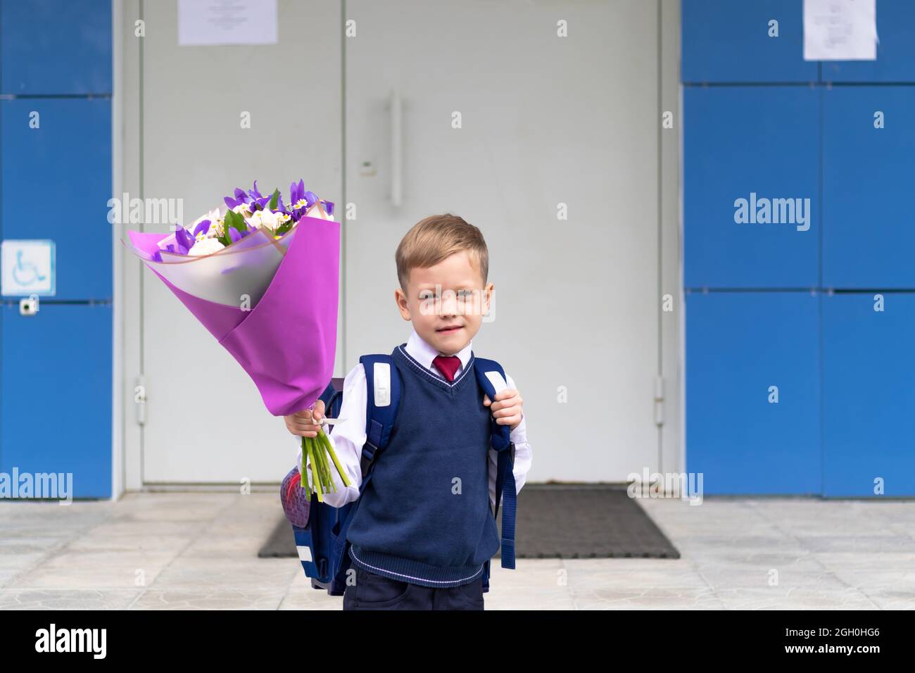 Ein süßer Erstklässler-Junge in Schuluniform mit einem schönen Blumenstrauß im Schulhof nach dem Feiertag am 1. September. Sachkenntniss Stockfoto