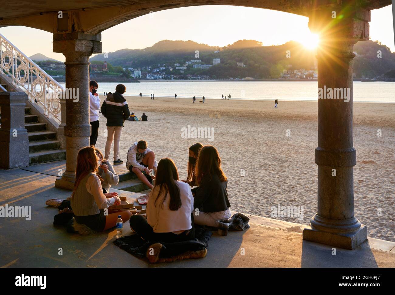 Eine Gruppe junger Freunde, die im Kreis sitzen und den Sonnenuntergang am Strand La Concha genießen, Donostia's Juwel par excellence, Wahrzeichen der Stadt, unvergleichlich Stockfoto