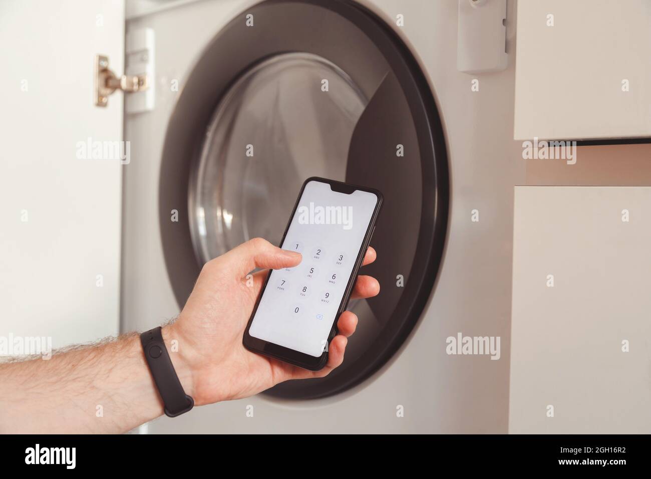 Mann benutzt Mobiltelefon, um die Waschmaschine zu steuern und zu starten. Smart-House-Systemgeräte. Stockfoto
