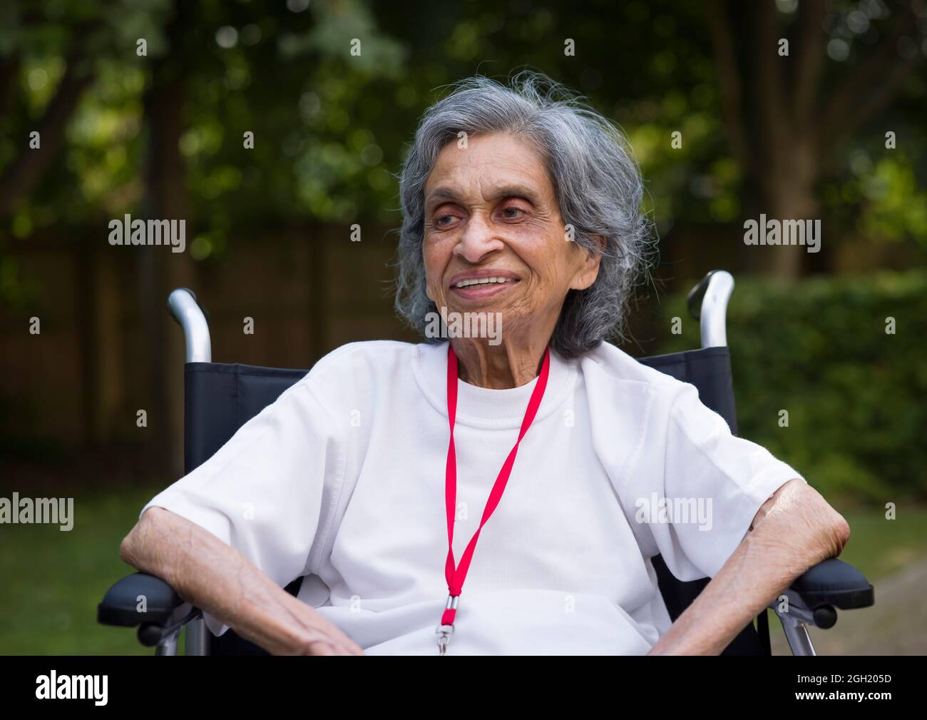 Alte indische Frau, die im Rollstuhl sitzt und lächelt. Im Sommer in Großbritannien im Freien in einem Park oder Garten Stockfoto