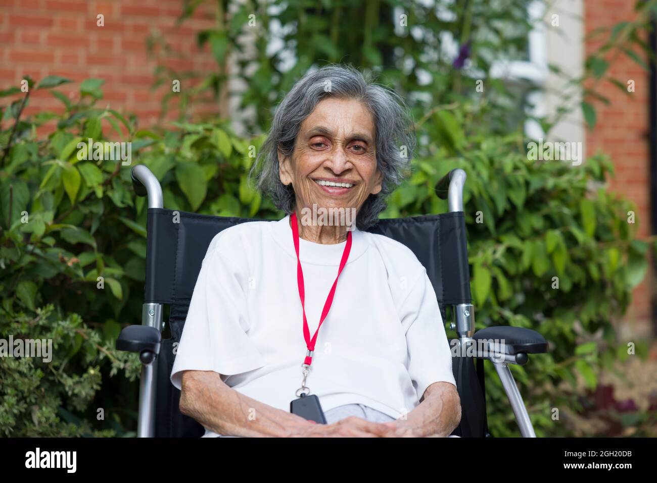 Medizin - Alte asiatische indische Frau, die lächelnd in einem Rollstuhl in einem Pflegeheim, Pflegeheim oder Garten sitzt, Großbritannien. Zeigt die Gesundheitsversorgung und den Ruhestand älterer Menschen Stockfoto