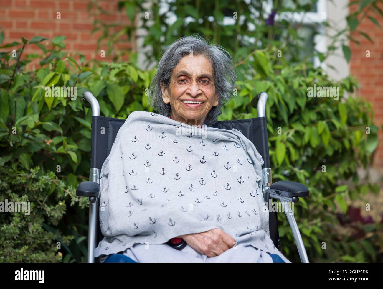 Ältere asiatische indische Frau, die lächelnd in einem Rollstuhl in einem Pflegeheim, Pflegeheim oder Garten sitzt, Großbritannien. Zeigt die Gesundheitsversorgung im Ruhestand und Alter Stockfoto