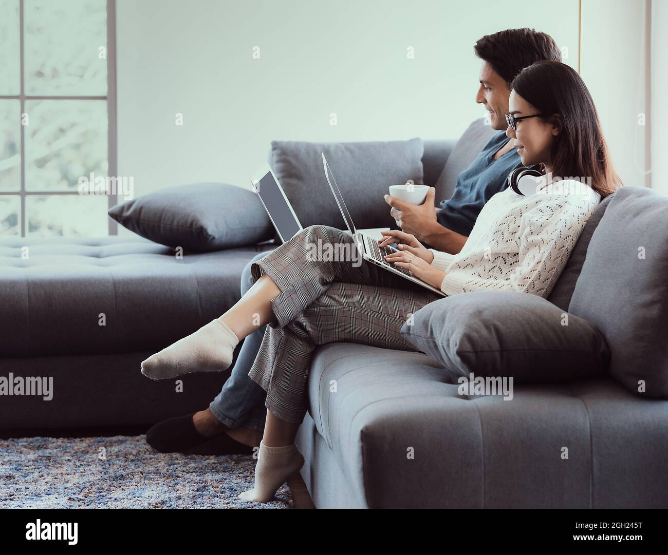 Mischen Sie Rennfamilienliebhaber, kaukasischen Mann und asiatische Frau, sitzen im Wohnzimmer arbeiten über Laptop-Computer auf dem Sofa in einfachen Entspannungsgesten. Konzept von Stockfoto