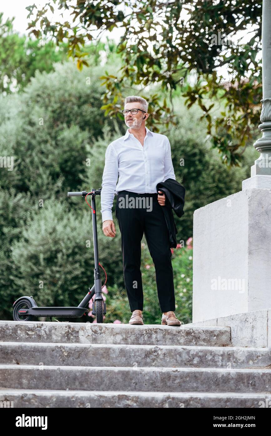 Porträt eines Geschäftsmannes mit Elektroroller, Jacke und Ohrhörern, während er Musik hört. Eleganter Manager mit guter Laune, der ein mobiles Gespräch führt Stockfoto