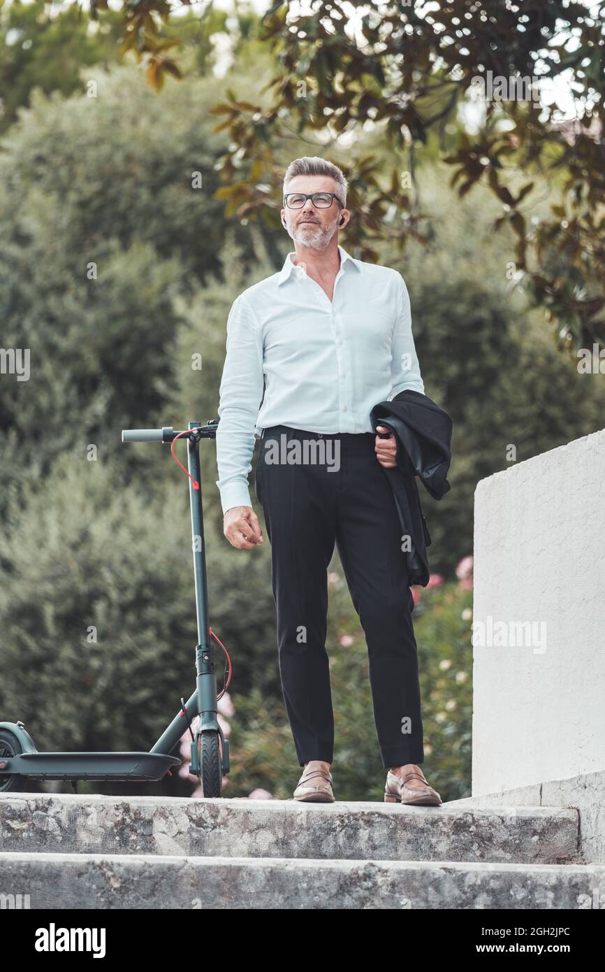 Porträt eines Geschäftsmannes mit Elektroroller, Jacke und Ohrhörern, während er Musik hört. Eleganter Manager mit guter Laune, der ein mobiles Gespräch führt Stockfoto