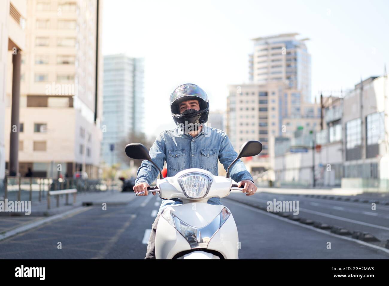 Die mittlere Aufnahme eines jungen Motorradfahrers hielt an einer Ampel in Barcelona an. Der Mann fährt mit seinem weißen Roller auf einer großen Allee voll durch die Stadt Stockfoto