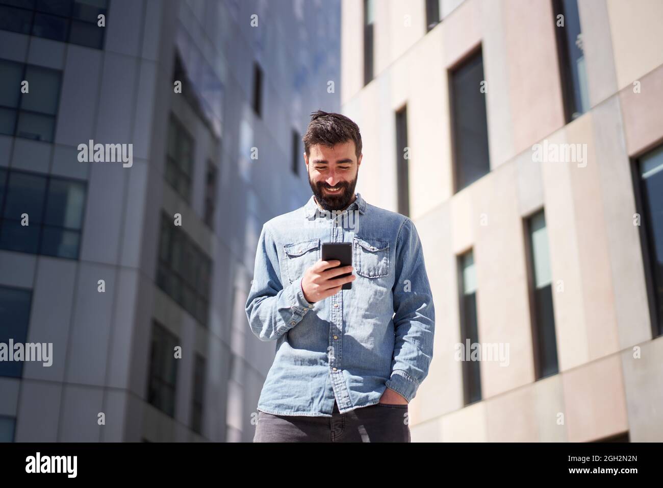 Junger Geschäftsmann im Denim-Hemd mit Smartphone in der Nähe von Bürogebäuden im Finanzzentrum der Stadt. Stockfoto