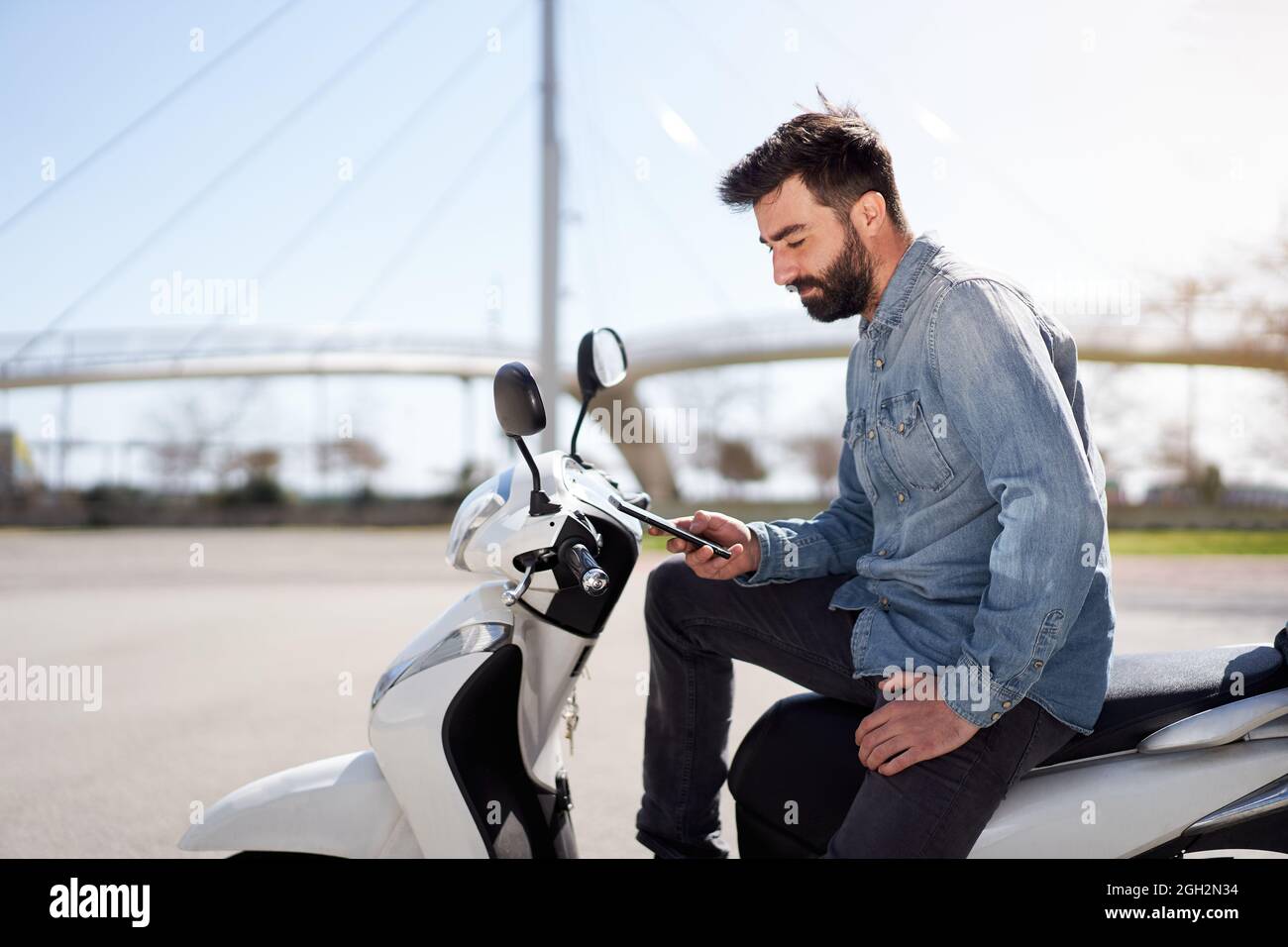 Junger Spanier, der Handy oder Smartphone benutzt und auf seinem Motorroller draußen in der Stadt sitzt. Geschäftsmann überprüft Arbeitsprobleme auf dem Handy. Stockfoto