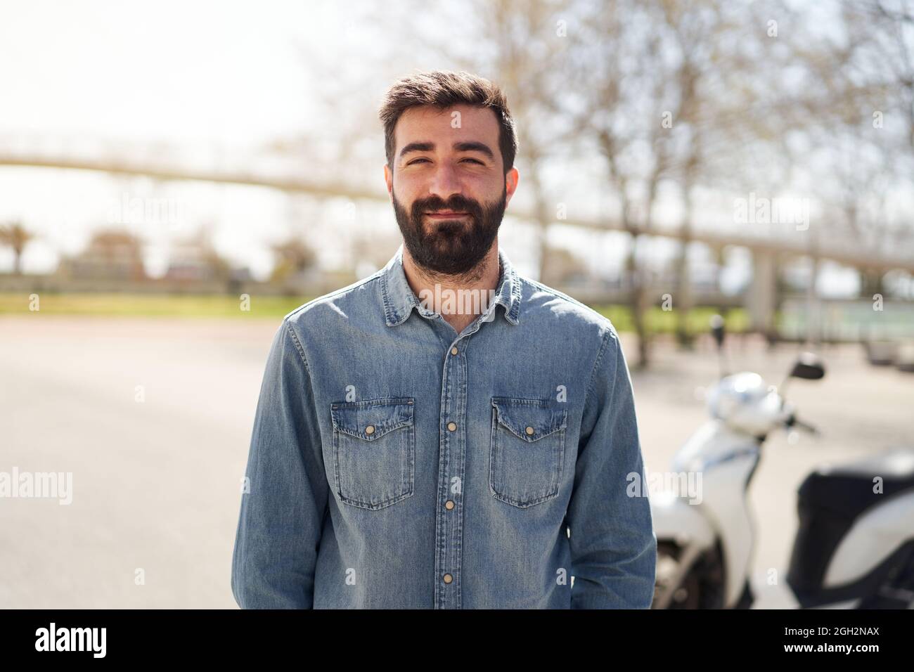 Porträt eines jungen Spanierers, der die Kamera anschaut. Der Mann mit fröhlichem Lächeln steht vor seinem Motorrad, das in der Stadt abgestellt wird. Stockfoto