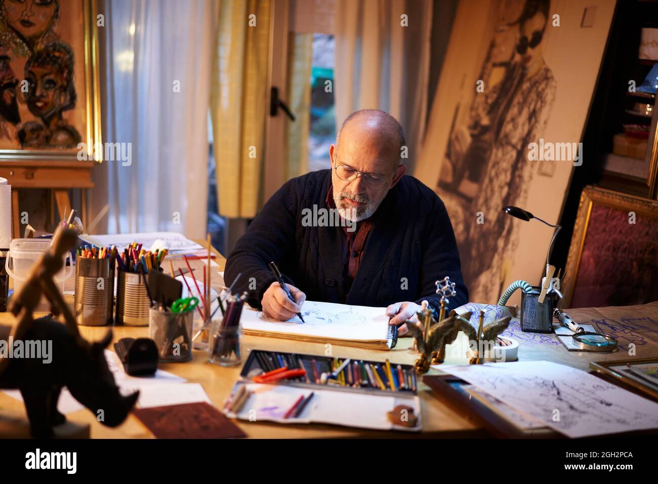 Künstler arbeitet im Studio. Älterer Mann malt ein Bild mit einer Lupe in einer kreativen und chaotischen Umgebung. Stockfoto