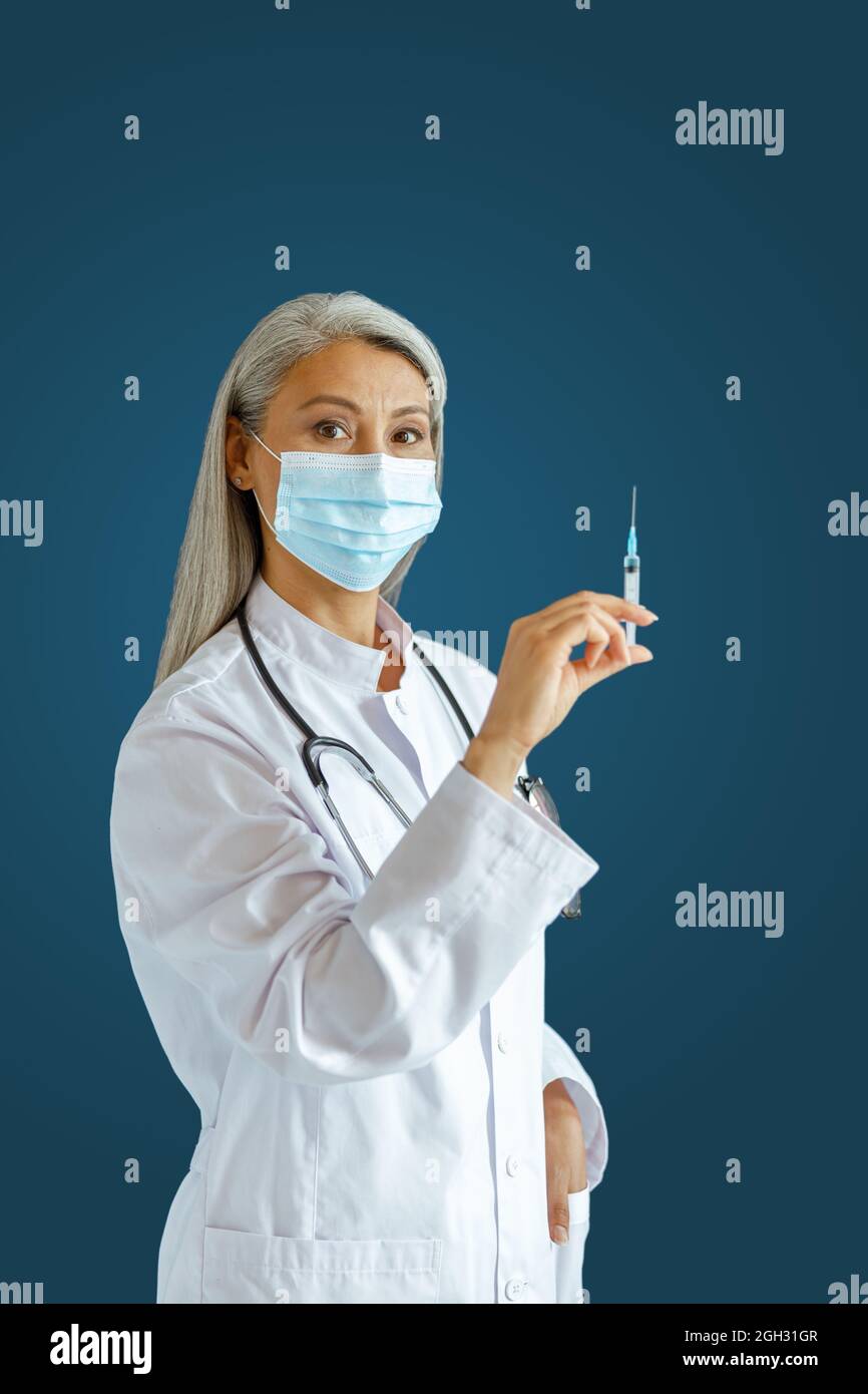 Silberhaarige asiatische Therapeutin mit Maske hält Spritze mit Medizin auf blauem Hintergrund Stockfoto