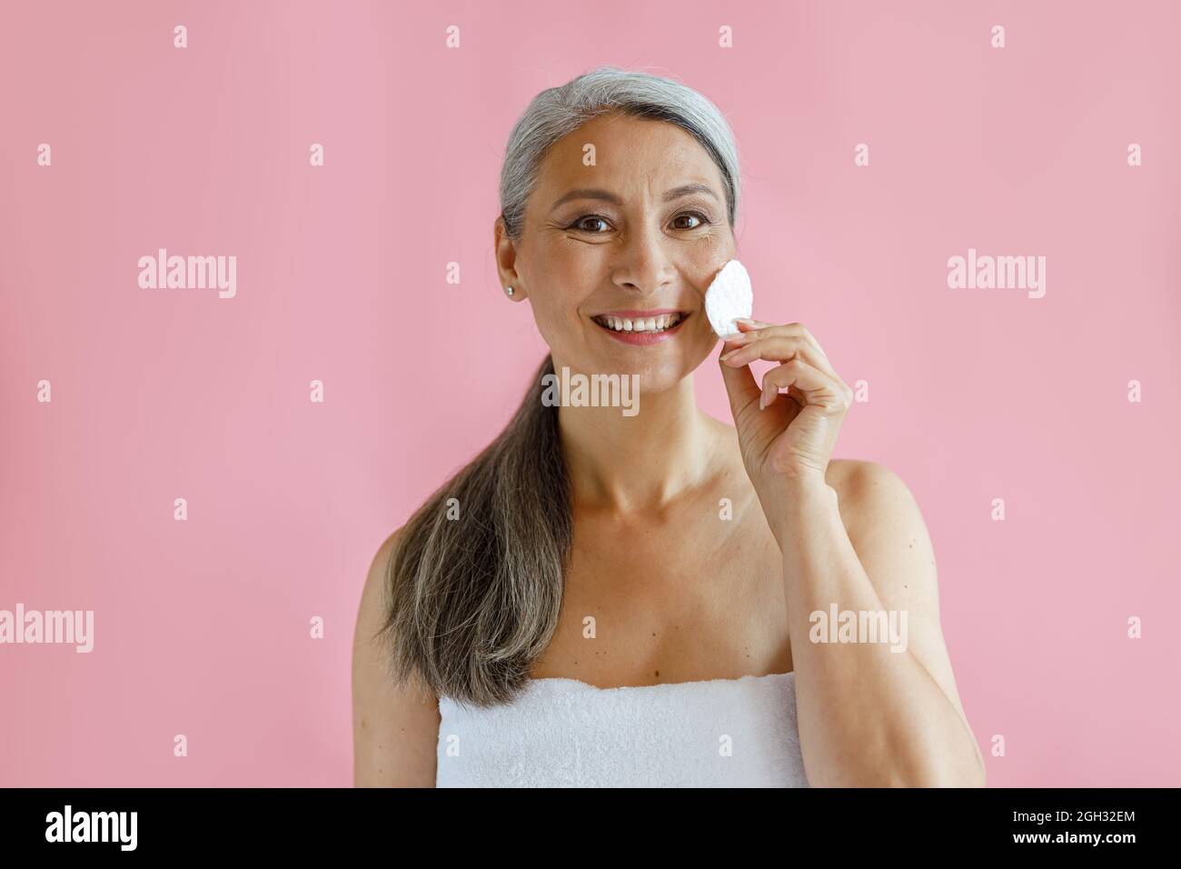 Lächelnd grauhaarige asiatische Frau reinigt Gesicht mit Wattepad stehen auf rosa Hintergrund Stockfoto