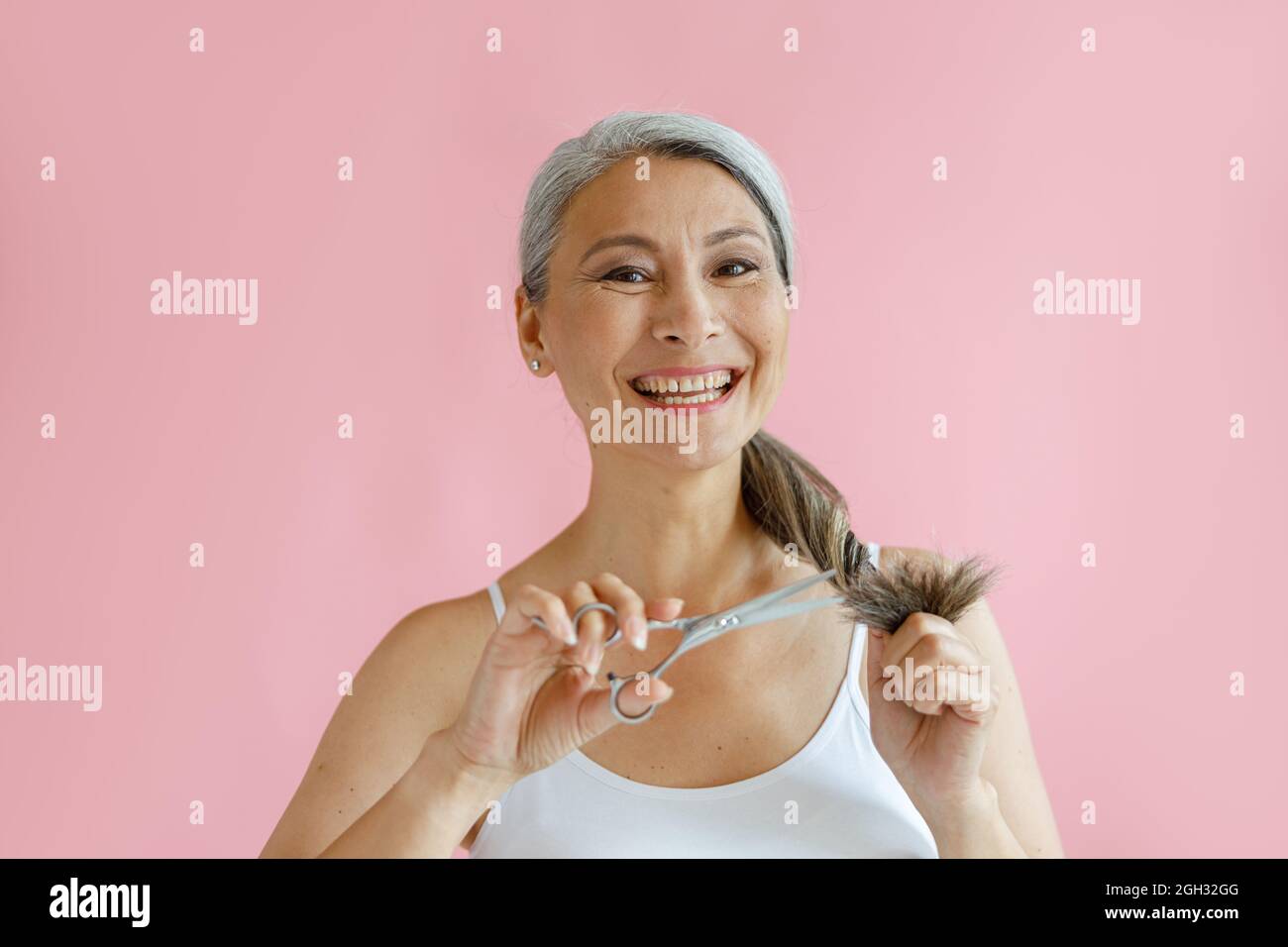 Lächelnde asiatische Dame mittleren Alters schneidet ausgefranste Enden von silbernen Haaren mit einer Schere auf rosa Hintergrund Stockfoto