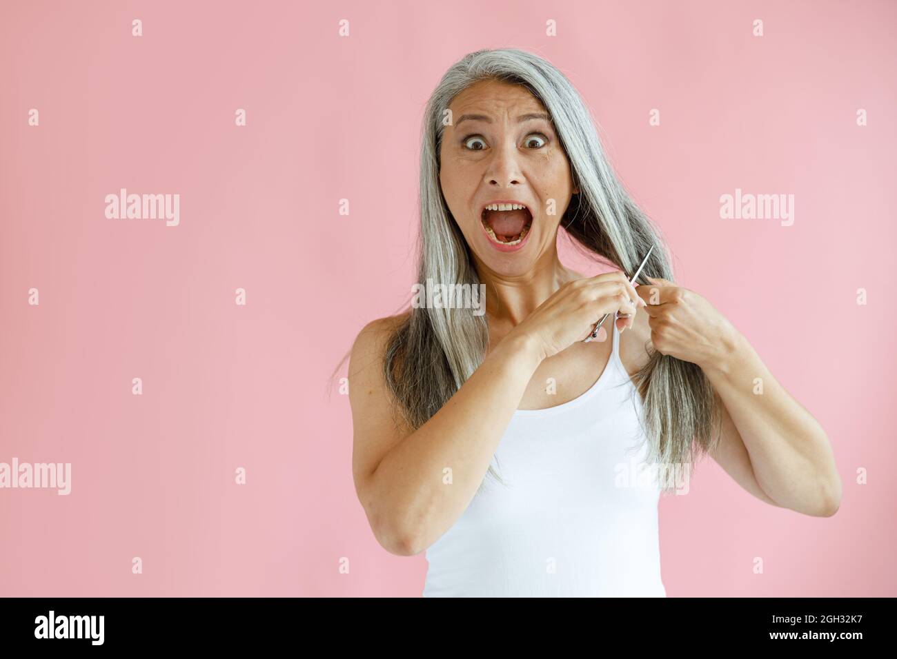 Aufgeregt mittleren Alters asiatische Frau schneidet lange silberne Haare mit scharfen Scheren auf rosa Hintergrund Stockfoto