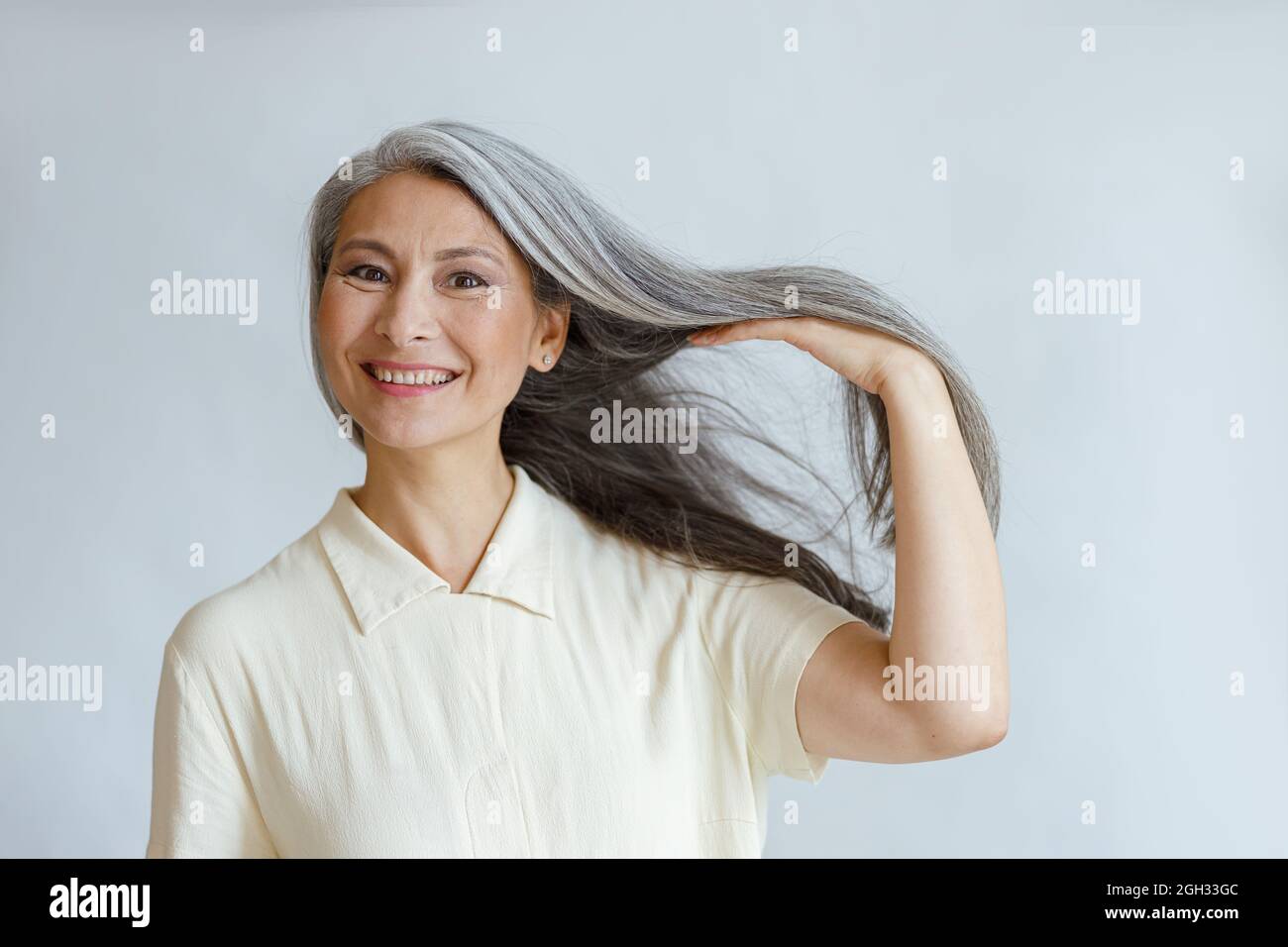 Glückliche asiatische Dame mittleren Alters zeigt natürliches silbernes Haar, das auf hellem Hintergrund posiert Stockfoto