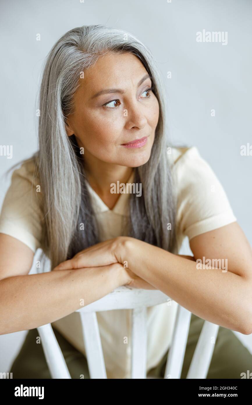 Eine hübsche Asiatin mittleren Alters mit geradem grauen Haar sitzt im Studio rückwärts auf einem Stuhl Stockfoto
