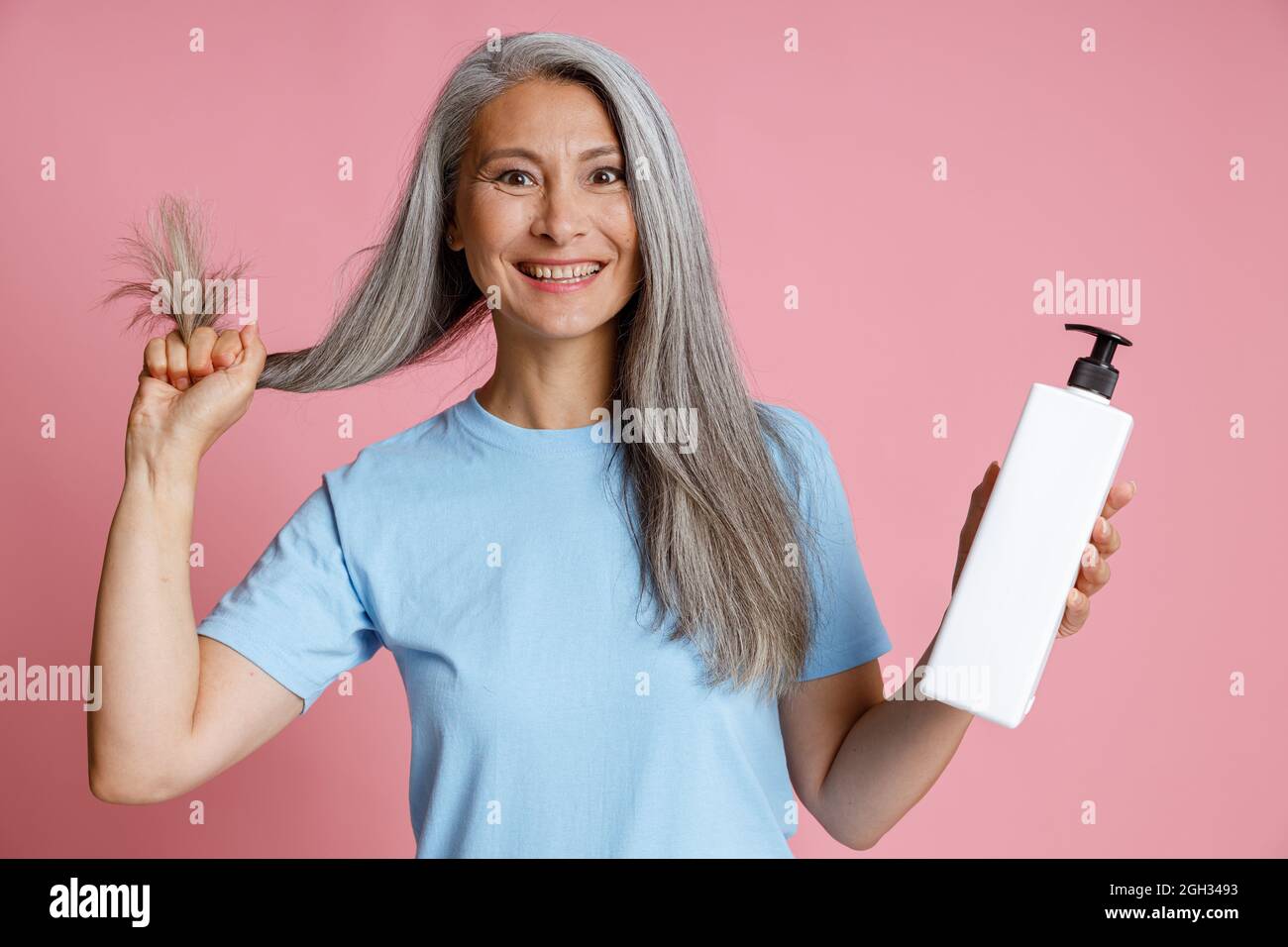 Asiatische Frau mittleren Alters zeigt gesunde silberne Haare und leere Spenderflasche auf rosa Hintergrund Stockfoto