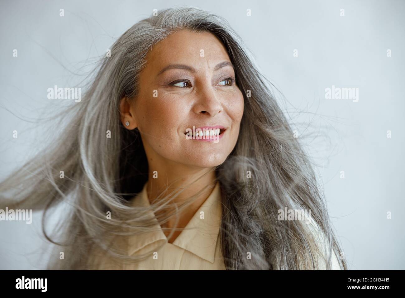 Glückliche asiatische Frau mittleren Alters mit fliegenden grauen Schlössern steht auf hellem Hintergrund Stockfoto
