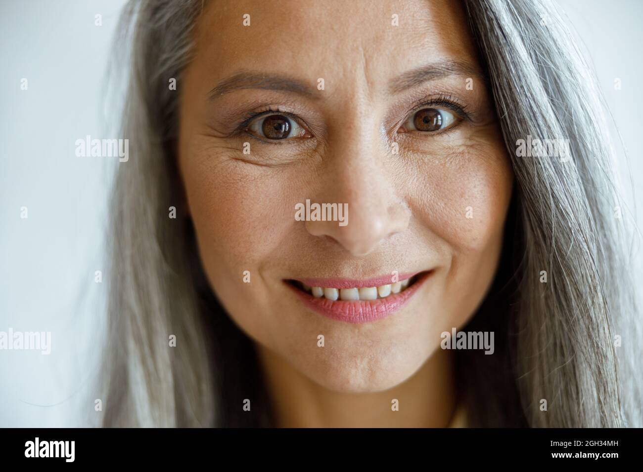 Fröhliche silberhaarige Dame mit Make-up schaut in die Kamera auf hellgrauem Hintergrund Stockfoto