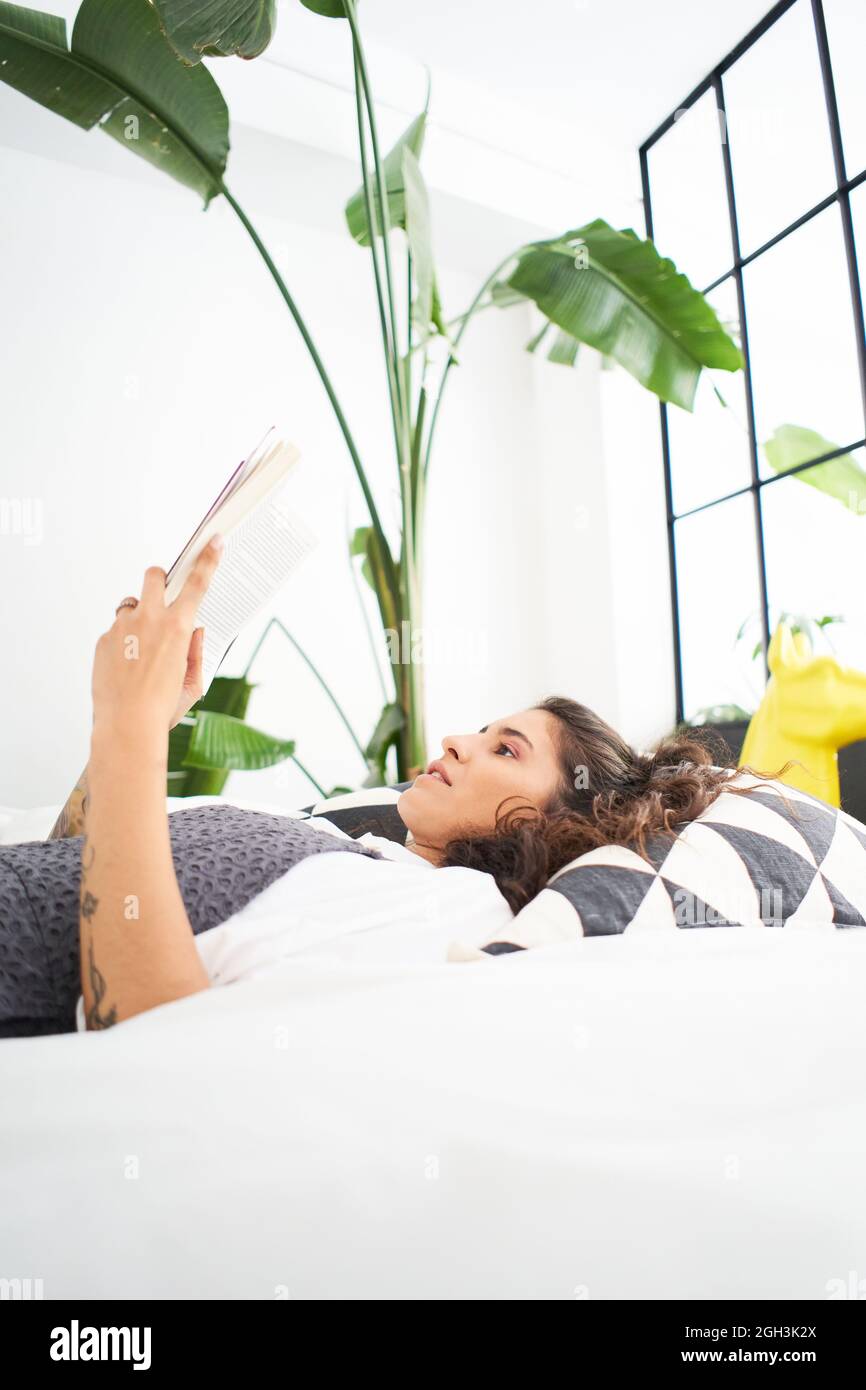 Vertikales Foto einer jungen Latina, die ein Buch liest, das auf dem Bett liegt. Menschen, die zu Hause studieren und lernen. Konzept von Freizeit und Kultur. Stockfoto