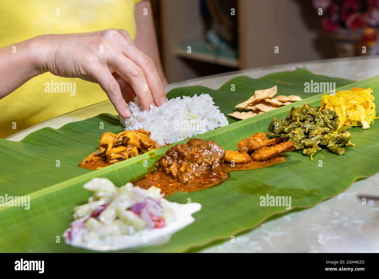 Person, die indischen Bananenblattreis aus Hammelfleisch-Paprikaschurry, Tintenfisch, Garnelen, Papadam und verschiedenen Gemüsesorten genießt Stockfoto