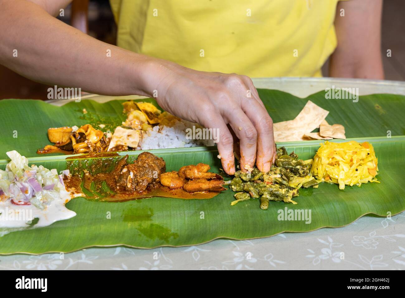 Person, die indischen Bananenblattreis aus Hammelfleisch-Paprikaschurry, Tintenfisch, Garnelen, Papadam und verschiedenen Gemüsesorten genießt Stockfoto