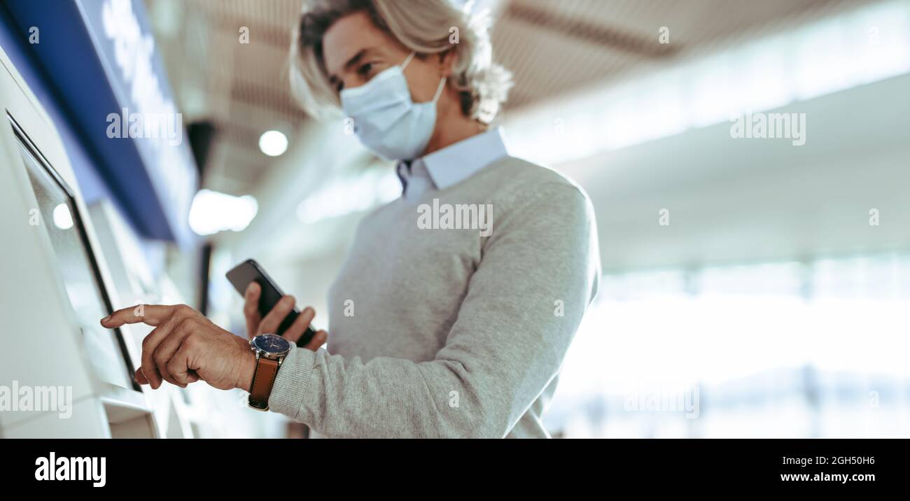 Geschäftsreisende, die am Flughafen mit einem Selbstbedienungsautomaten einchecken. Geschäftsmann, der eine Gesichtsschutzmaske trägt, checkte mit der automatischen Maschine bei einem ein Stockfoto