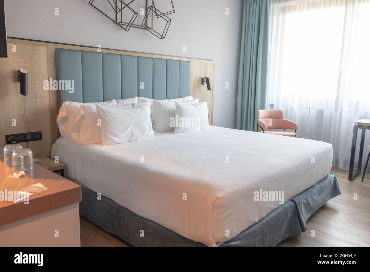 Helles und gemütliches, modernes Hotelzimmer mit komfortablem Kingsize-Bett, großem Fenster und blauem Kopfteil. Stockfoto