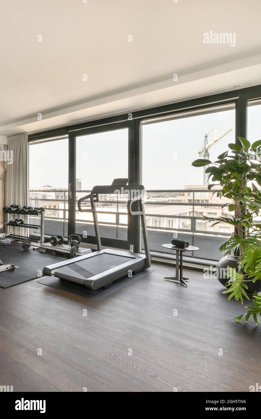 Die Inneneinrichtung der geräumigen Halle mit Laufband und Sportartikeln befindet sich vor einem großen Panoramafenster mit Blick auf die Stadt in einem modernen, luxuriösen Apartment Stockfoto