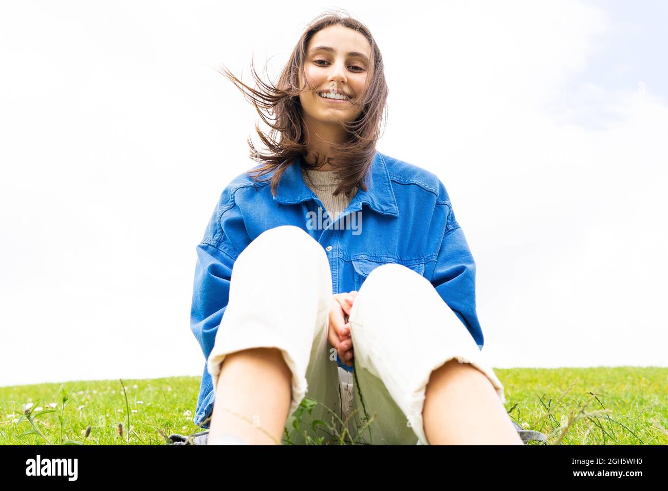 Ein niedriger Winkel von glücklicher Frau, die an windigen Tagen auf einem Hügel in der Natur sitzt, während sie wegschaut Stockfoto