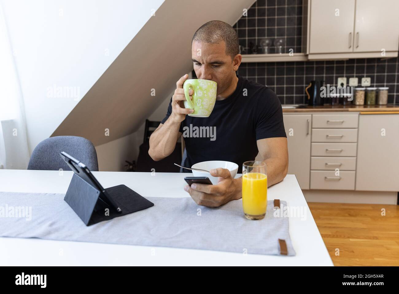 Erwachsener, der ein heißes Getränk aus dem Becher trinkt und im Mobiltelefon stöbert, während er am Tisch in der Küche sitzt und morgens zu Hause frühstücken kann Stockfoto